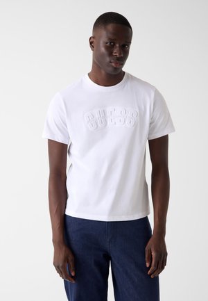 Młody mężczyzna w białej, tłoczonej koszulce "GUESS" i ciemnoniebieskich dżinsach, stojący na jednolitym białym tle.