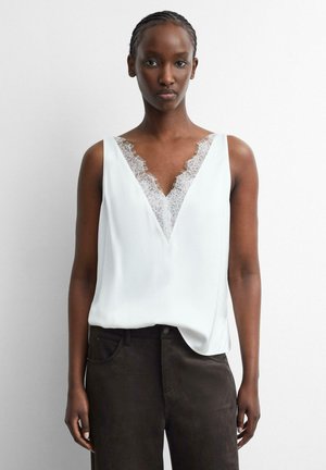 Donna che indossa una camicetta bianca senza maniche con rifiniture in pizzo lungo una profonda scollatura a V e pantaloni scuri a vita alta, in piedi davanti a uno sfondo semplice.