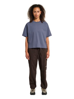 Kvinde med krøllet hår står med front mod, iført en løs blå T-shirt, mørkebrune bukser og beige vandresko.