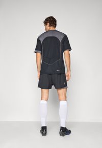Man in zwart en grijs sportoutfit, met witte knielange sokken en zwarte voetbalschoenen, staand met zijn gezicht naar een effen lichtgrijze achtergrond.