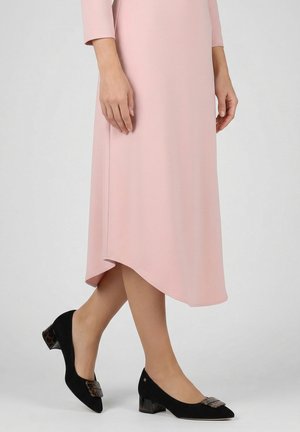 Frau trägt ein knielanges hellrosa Kleid und schwarze flache Schuhe mit dekorativer quadratischer Schnalle, sie steht auf einem grauen Boden.