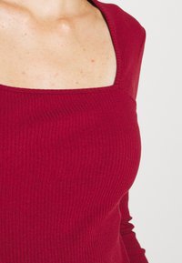 Burgunderfarbener Rippoberteil mit quadratischem Ausschnitt und langen Ärmeln, aus einem strukturierten, dehnbaren Stoff.
