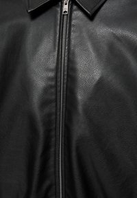 Veste en cuir noir avec une texture visible, une fermeture éclair métallique à l'avant partiellement fermée et un col replié vers le bas en haut.