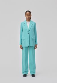 Een turquoise twee-delige pak bevat een op maat gemaakte blazer met een enkele knoop en twee zakken, gecombineerd met wijde broekspijpen en zwarte schoenen.