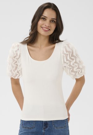 Femeie purtând un top alb cu mâneci bufante transparente și tivuri, zâmbind cu mâinile la spate, îmbrăcată în blugi albaștri.