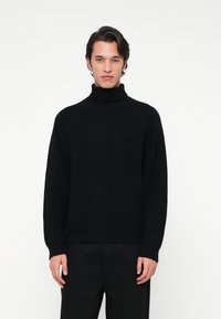 Jeune homme faisant face, portant un pull col roulé noir à côtes et un pantalon noir, sur fond blanc uni.