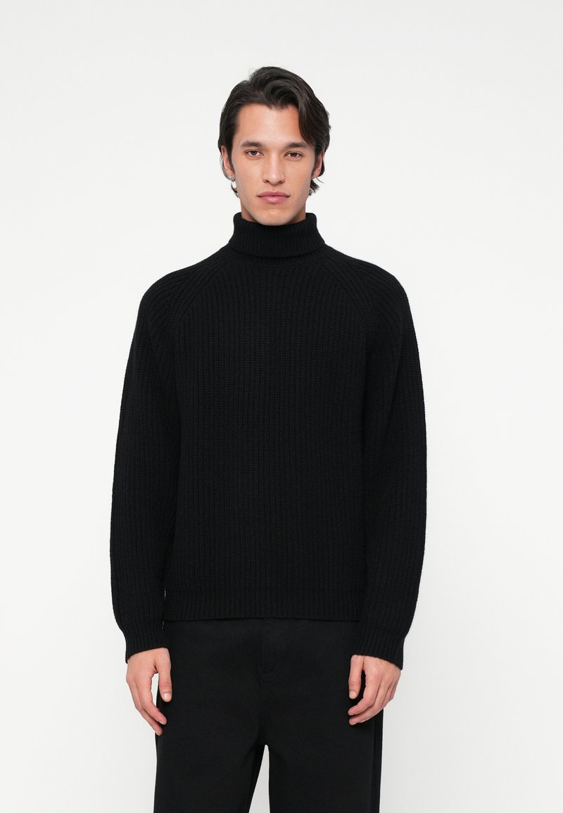 Jeune homme faisant face, portant un pull col roulé noir à côtes et un pantalon noir, sur fond blanc uni.