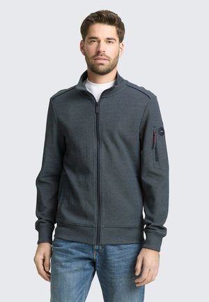 Mężczyzna ubrany w ciemną kurtkę na zamek z małą kieszenią na zamek na lewym rękawie i niebieskie dżinsy, stojący na tle jasnego, gładkiego tła.