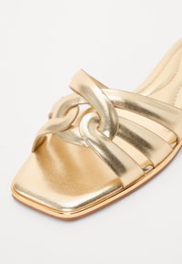 Gullfarget metallisk sandal med firkantet tå og sammenflettede stropper, med en jevn tekstur og en slank, flat såle.