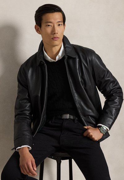 Homme assis sur un tabouret portant une veste en cuir noire, un pull noir sur une chemise blanche, un pantalon noir et une montre-bracelet avec un cadran vert.