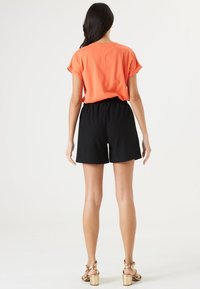 Pomarańczowa koszula z krótkim rękawem zestawiona z czarnymi, teksturowanymi szortami z elastycznym pasem. Modelka nosi złote sandały na obcasie.