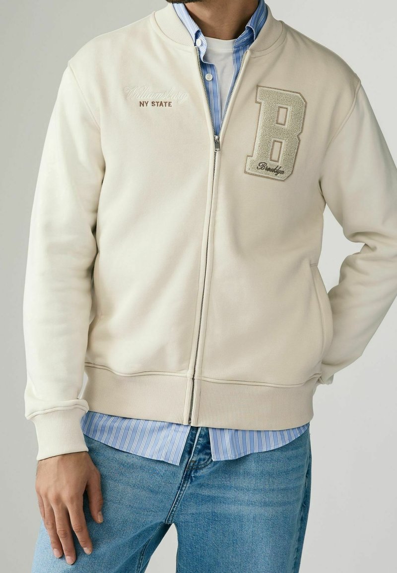 Giacca bomber beige con chiusura a zip, polsini e orlo a costine. Presenta un ricamo con la lettera "B" e il testo "Williamsburg NY STATE" sul petto.