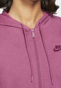 Roze zip-up hoodie van zacht materiaal met een textuurafwerking, voorzien van een capuchon met trekkoord en een geborduurd Nike-logo op de borst.
