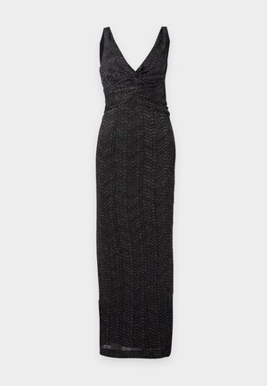 Un mannequin se tient devant un fond uni, vêtu d’une robe noire sans manches, longue jusqu’au sol, avec un décolleté en V, une taille froncée et une fente latérale, ainsi que des sandales à talons.