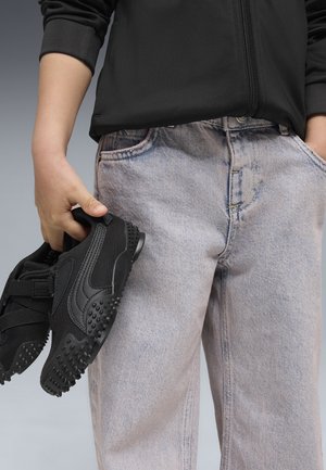 Osoba ubrana w jasne dżinsy i czarną kurtkę, trzymająca w jednej ręce parę czarnych sneakersów z chropowatą podeszwą.