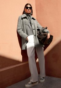 Vrouw in een grijze jas, donkergrijze coltrui, witte broek, sneakers, die een grote zwarte tas vasthoudt en tegen een perzikkleurige muur in het zonlicht staat.