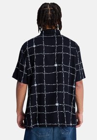 Κοντομάνικο πουκάμισο από μαύρο ύφασμα με λευκό σχέδιο σε πλέγμα. Διαθέτει άνετη εφαρμογή και γιακά.