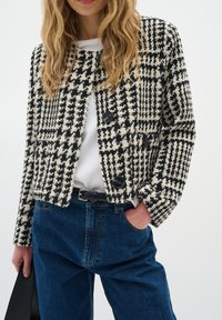 Černobílá kostkovaná zkrácená bunda vyrobená z texturované tkaniny, s dvěma velkými knoflíky, spárovaná s modrými džíny s vysokým pasem.