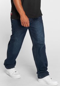 Donkerblauwe spijkerbroek met een relaxte pasvorm, omgeslagen pijpen en een gladde textuur, gecombineerd met witte sportieve sneakers.