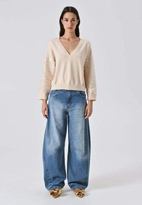 Maglione beige con scollo a V e maniche impreziosite da paillettes, abbinato a jeans a gamba larga color blu sbiadito e scarpe oro con punta appuntita. Sfondo semplice.