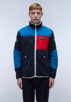 Giacca in pile con parte superiore blu, tasca sul petto rossa e parte inferiore blu scuro. Chiusura frontale con zip, rifiniture bianche e tessuto strutturato.