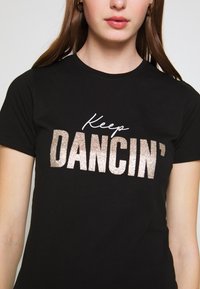 Camiseta negra de algodón con el texto "Keep DANCIN'" en fuentes brillantes plateadas y blancas. Manga corta y cuello redondo.
