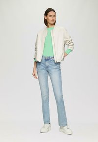 Veste bomber légère crème sur un pull vert, associée à un jean bleu et des baskets blanches à plateforme. Design simple, coupe décontractée.
