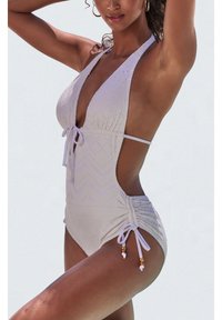 Femme portant un maillot de bain une pièce violet clair avec des découpes latérales, un laçage devant et des côtés froncés ornés de perles décoratives, posant les bras levés.