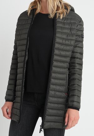 Femme portant une longue doudoune matelassée vert foncé sur une chemise noire et un pantalon foncé, debout devant un fond uni.