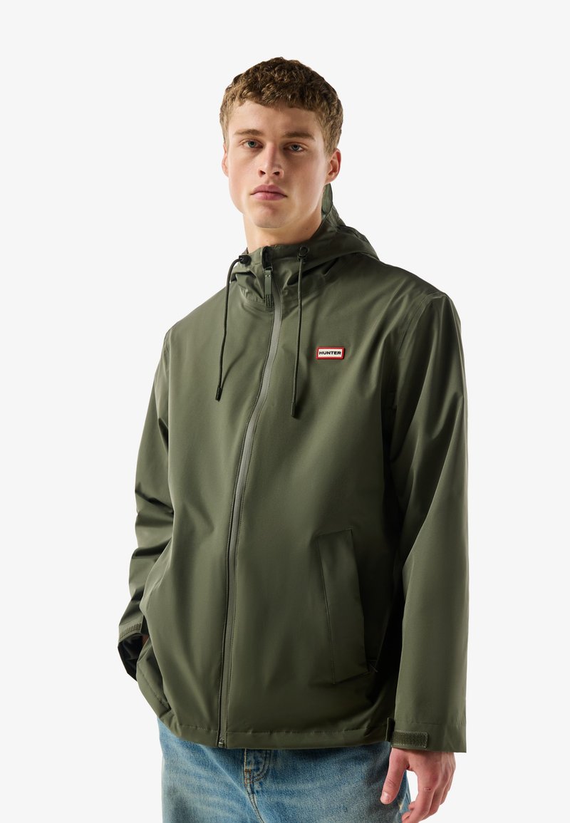 Junger Mann trägt eine grüne Regenjacke mit Reißverschluss, Kapuze und Logo auf der Brust, steht mit teilweise in den Taschen steckenden Händen vor weißem Hintergrund.