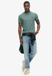 Groene heather crewneck T-shirt, blauwe jeans, een geruit flanel overhemd geknoopt op de taille en beige sneakers, benadrukken een casual stijl en gelaagde texturen.
