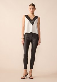 Top blanco sin mangas con escote en V de encaje negro, combinado con pantalones ajustados de cuero negro y sandalias de tacón negro. Textura suave en general.