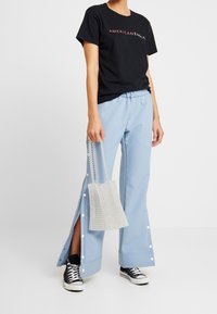 Svart T-shirt med texten "AMERICAN EAGLE", ljusblå byxor med tryckknappar på sidan, och en transparent handväska prydd med pärlor. Svarta sneakers.