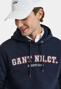 Marineblauer Kapuzenpullover mit dem Text "GANT NIL. CT. SPORTS CLUB" in Weiß und Rot, ausgestattet mit einer Zugkordelkapuze und weichem Material. Weißer Hut mit "Gant"-Logo.