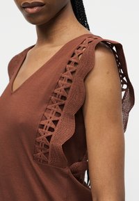 Κοντινό πλάνο ενός ατόμου που φοράει καφέ αμάνικο τοπ με V-λαιμόκοψη και διακοσμητική πλεκτή δαντέλα στον ώμο και την μασχάλη.