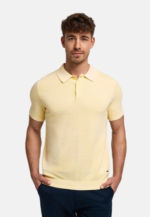 Homme portant un polo jaune pâle à manches courtes avec boutons, pantalon bleu marine, debout les mains dans les poches devant un fond uni clair.