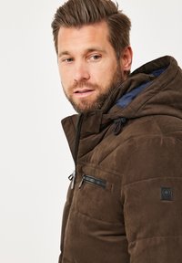 Braune gepolsterte Jacke mit Kapuze, die eine glatte Textur aufweist, eine mit Reißverschluss verschlossene Brusttasche hat und ein Logopatch am Ärmel trägt. Abgerundet mit einem marineblauen Innenfutter.