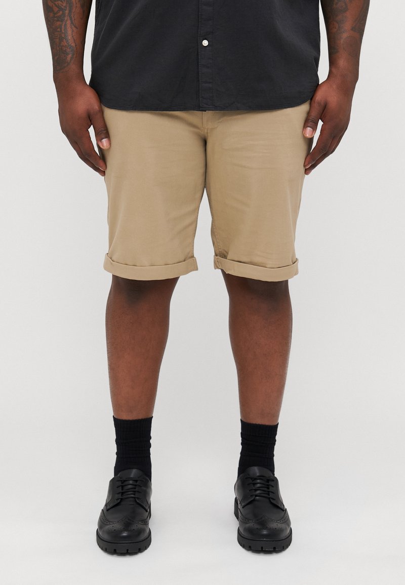 Khaki-Shorts mit umgeschlagenen Säumen, getragen mit schwarzen Socken und schwarzen Schuhen. Das Hemd ist dunkel und zugeknöpft. Glatte Textiloberflächen sind sichtbar.