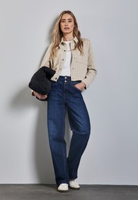 Ljust beige randig skjorta, vit t-shirt, mörkblå vida jeans och krämfärgade sneakers. Modellen håller en svart puffjacka.