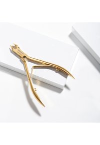 Złote szczypce do skórek ze stali nierdzewnej z wygiętymi uchwytami spoczywające na białych prostokątnych blokach na białym tle.