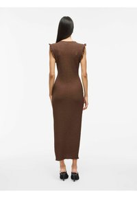 Długa, dopasowana brązowa sukienka z fakturowanej tkaniny, z falbaniastymi akcentami na ramionach i prostym dołem. Modelka nosi czarne szpilki.
