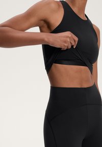 Top sportivo nero che viene sollevato, rivelando un bralette nero sottostante. Materiale liscio ed elastico con cuciture minime.