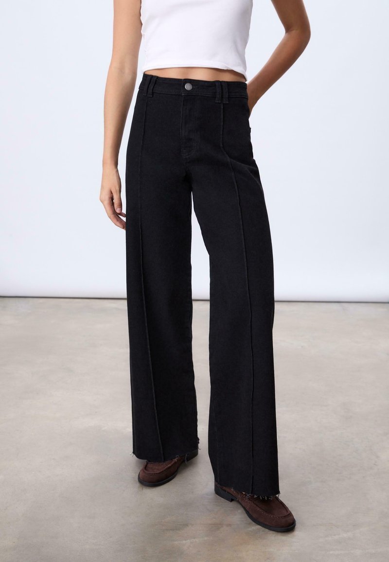 Femme portant un jean noir taille haute à jambes larges avec des ourlets bruts, des chaussures marron, et un top blanc court sans manches, debout sur un sol beige.