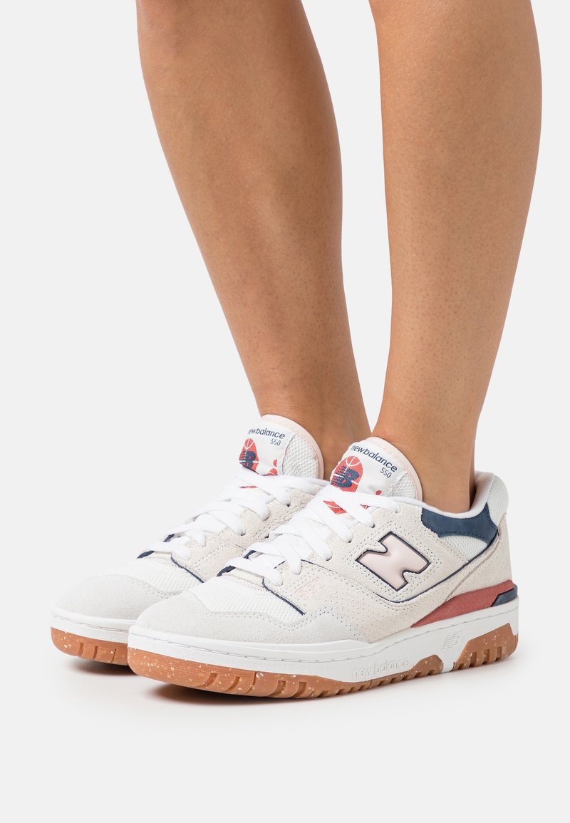 Baskets New Balance 550 blanches et bleu marine avec semelles en gomme portées pieds nus sur un fond clair uni.