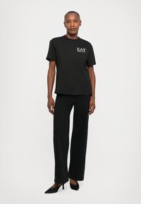 Kvinne iført en svart EA7 Emporio Armani t-skjorte, vide svarte bukser og svarte hæler, stående mot en hvit bakgrunn.