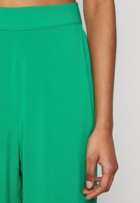 Zelené široké kalhoty s vysokým elastickým pasem; hladká textura a minimální švy viditelné na tkanině.