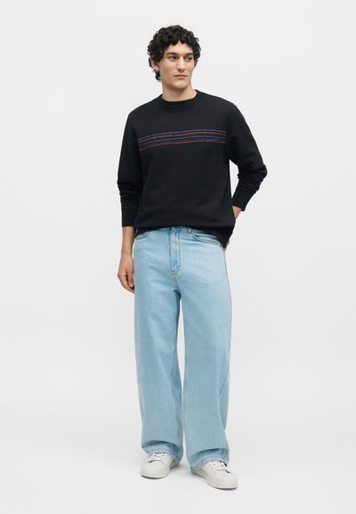 Άνδρας με σγουρά μαλλιά που φοράει μαύρο μπλουζάκι μακρυμάνικο με πολύχρωμες ρίγες, ανοιχτό μπλε παντελόνι με φαρδιά μπατζάκια και λευκά αθλητικά παπούτσια.