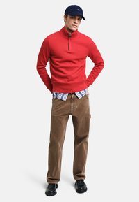 Roter Sweatshirt mit Reißverschluss, kombiniert mit einem blau-weiß gestreiften Hemd mit Kragen, braunen Cordhosen und schwarzen Schuhen. Der Hut ist dunkelblau.