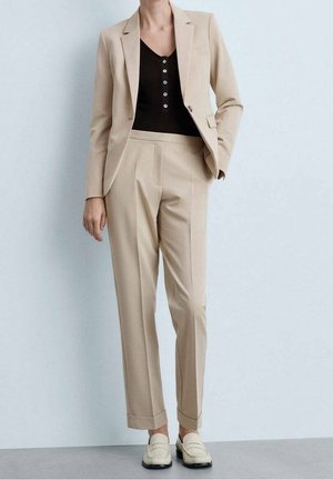 Femme portant un costume beige ajusté avec un blazer à un seul bouton, un haut noir boutonné et des mocassins crème, se tenant devant un fond clair uni.