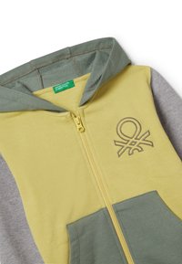 Sudadera con cremallera en amarillo, verde y gris. Presenta una textura suave, capucha y un diseño bordado. Incluye un cierre con cremallera y bolsillos frontales.